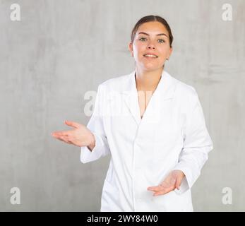 Dottore in camice bianco sorridente alla macchina fotografica mentre si presenta con la mano e punta con il dito Foto Stock