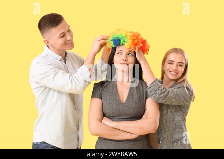 Uomini d'affari che mettono la parrucca da clown sul loro collega su sfondo giallo. Festa del Fool's Day di aprile Foto Stock