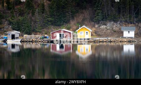 Quattro piccoli capannoni di barche seduti sul bordo delle rive si riflettono sulle acque calme di fronte a loro. Foto Stock