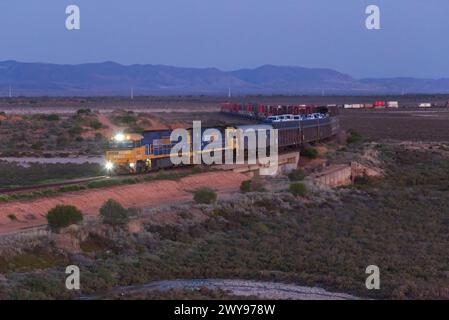 Treno merci che si muove attraverso un paesaggio al crepuscolo con montagne sullo sfondo e un cielo limpido. Foto Stock