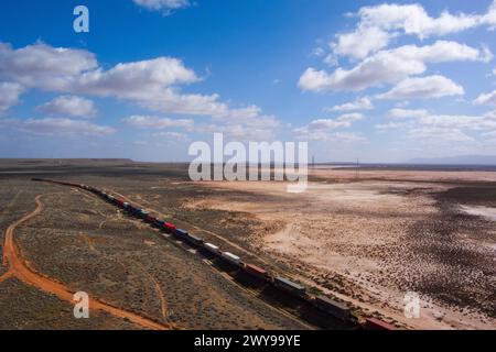 Vista aerea di un lungo treno merci che si muove attraverso un vasto e arido paesaggio desertico sotto un cielo blu Foto Stock