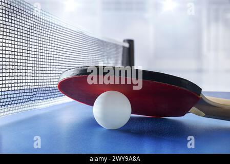 Pagaia da ping pong appoggiata su una palla bianca su un tavolo da gioco blu accanto alla rete di gioco con padiglione sportivo con illuminazione sullo sfondo. Vista frontale. Foto Stock