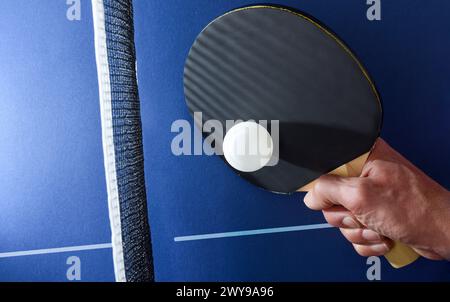Dettaglio della mano con pala da ping pong che colpisce una palla bianca accanto alla rete su un tavolo da gioco blu. Vista dall'alto. Foto Stock