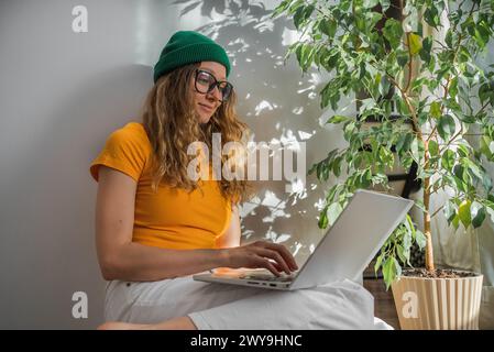 Una donna concentrata lavora dal suo portatile in un ambiente domestico, accompagnato dalla rilassante presenza di una lussureggiante pianta interna e dal gioco di luce attraverso le foglie sulla parete Foto Stock