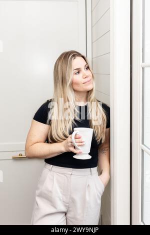 Donna che tiene in mano una tazza da caffè, perduta nel pensiero in un interno elegante Foto Stock