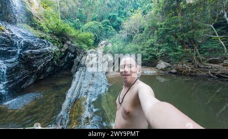 Un uomo fa un selfie con una pittoresca cascata in un ambiente lussureggiante della foresta. Foto Stock