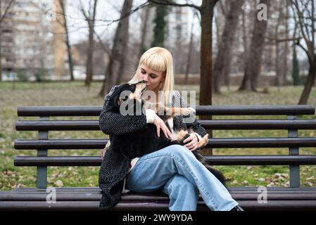 La giovane generazione Z hipster ragazza seduta in un parco su una panchina con il suo cane adottato, che ha salvato dalla strada dalla fame, e gli dà amore Foto Stock