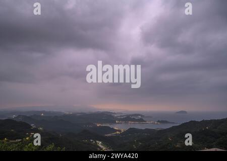 Una pittoresca scena serale che mostra una città costiera illuminata sotto un cielo scuro, con colline ondulate e l'oceano in vista Foto Stock