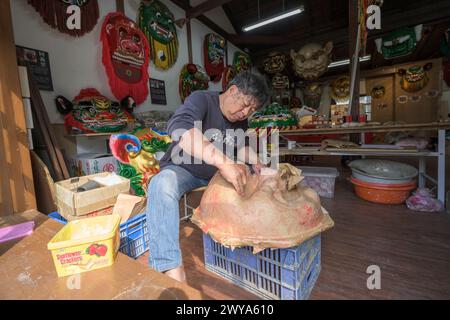 Un artigiano sta realizzando a mano una maschera cinese in un laboratorio illuminato dal sole circondato da maschere e decorazioni colorate Foto Stock