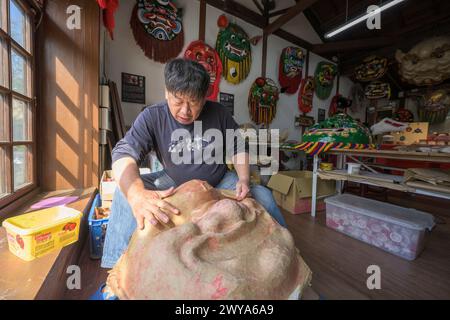 Un artigiano sta realizzando a mano una maschera cinese in un laboratorio illuminato dal sole circondato da maschere e decorazioni colorate Foto Stock