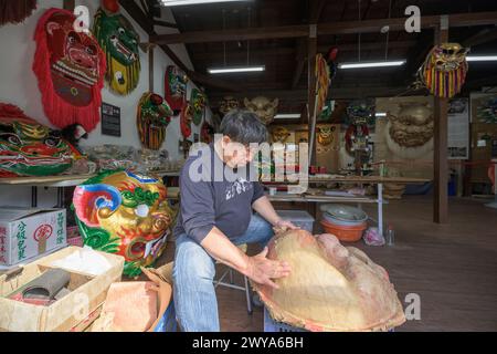 Un artigiano sta realizzando a mano una maschera cinese in un laboratorio illuminato dal sole circondato da maschere e decorazioni colorate Foto Stock