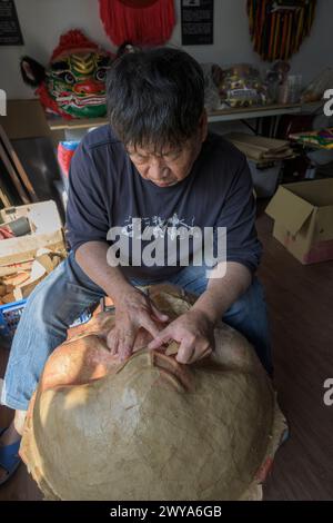 Un artigiano sta realizzando a mano una maschera cinese in un laboratorio illuminato dal sole circondato da maschere e decorazioni colorate Foto Stock