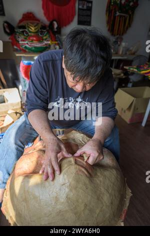 Un artigiano sta realizzando a mano una maschera cinese in un laboratorio illuminato dal sole circondato da maschere e decorazioni colorate Foto Stock