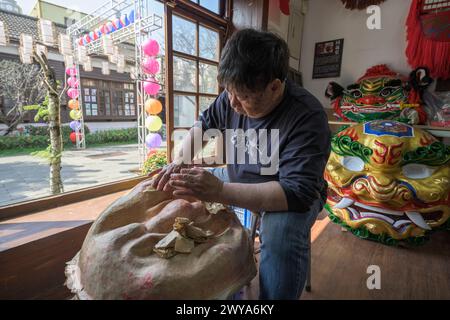 Un artigiano sta realizzando a mano una maschera cinese in un laboratorio illuminato dal sole circondato da maschere e decorazioni colorate Foto Stock