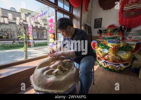 Un artigiano sta realizzando a mano una maschera cinese in un laboratorio illuminato dal sole circondato da maschere e decorazioni colorate Foto Stock