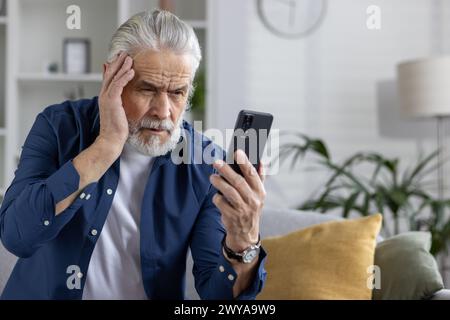 Un anziano preoccupato a casa che guarda il suo telefono, mostra emozioni di ricevere notizie sfortunate o cattive, tenendo testa in difficoltà. Foto Stock