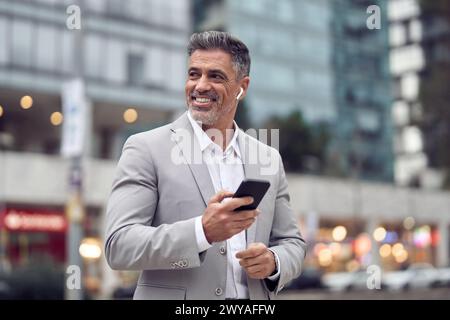 Uomo d'affari felice e maturo con auricolari che parla al telefono camminando lungo le strade trafficate della città. Foto Stock
