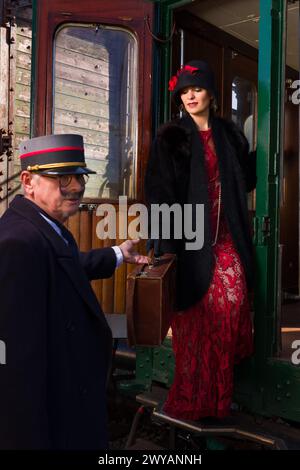 Attraente giovane donna in abito flapper rosso 1920s e cappello di cloche che viaggia con un antico treno a vapore Foto Stock