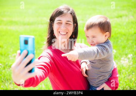 La madre porta il suo bambino di due anni tra le braccia mentre fanno un selfie vicino a un prato verde primaverile. I loro volti mostrano un grande sorriso di felicità. Foto Stock