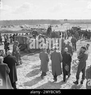 I detenuti a Bergen-Belsen osservano le guardie delle SS che caricano i corpi su camion mentre gli ufficiali dell'esercito britannico scortano i visitatori civili durante la liberazione dell'aprile 1945. Foto Stock
