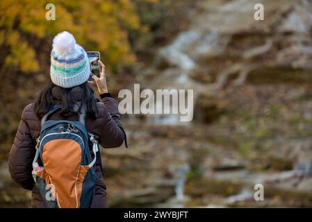 donna che scatta una foto di una cascata in autunno con il cambio delle foglie sul suo cellulare (escursionista con zaino e giacca, cappello scatta una foto al telefono cellulare o Foto Stock