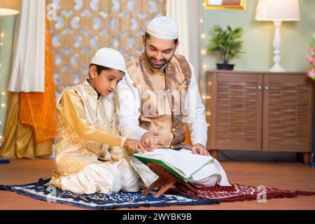 Padre musulmano indiano di supporto che aiuta il bambino a leggere il corano mentre fa la namza durante il festival ramdan a casa - concetto di festival, famiglia di sostegno Foto Stock