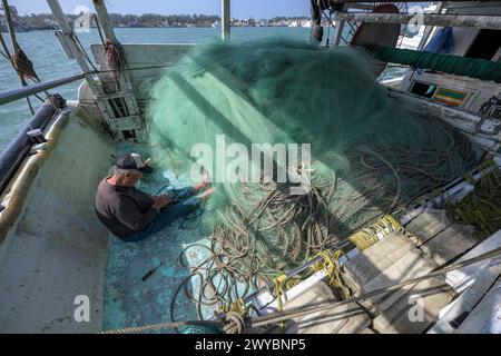 Un pescatore ripara diligentemente le reti da pesca su un ponte di barche, mostrando la vita quotidiana del lavoro marittimo Foto Stock