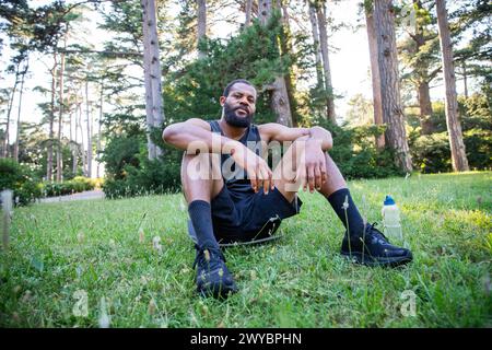 Un uomo sportivo siede sull'erba in un parco, indossando pantaloncini corti e canotta. Ha una bottiglia d'acqua accanto a lui Foto Stock