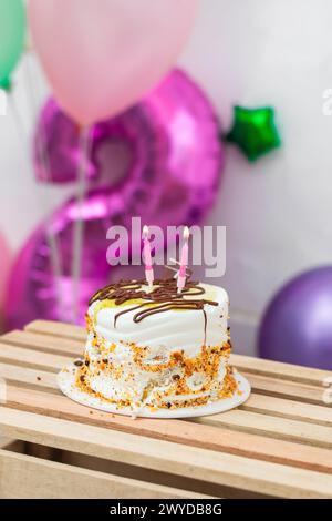 piccola torta di compleanno con due candele accese, sopra un tavolo di legno con palloncini sullo sfondo Foto Stock