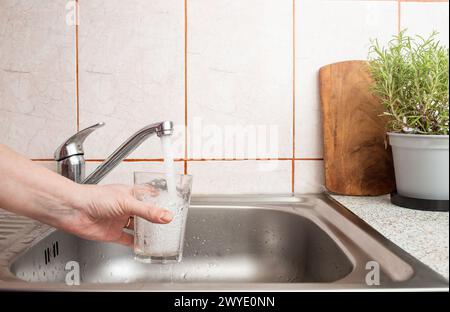 Donna che tiene a mano un bicchiere trasparente sotto un flusso di acqua corrente fresca da un rubinetto del lavandino della cucina, fuoco morbido Foto Stock