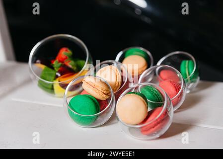 Una deliziosa disposizione di macaron colorati e pezzi di frutta fresca in bicchieri di vetro trasparente, esposti su una superficie bianca per una presentazione appetitosa. Foto Stock