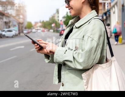 Una donna cerca un veicolo condiviso o a noleggio per strada utilizzando il suo smartphone. Una donna sta accanto alla strada in città usando il suo telefono per cercare Foto Stock