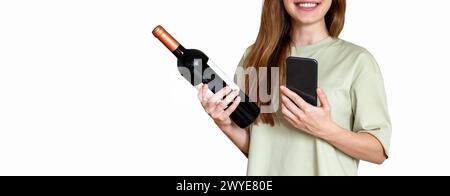 Una bella donna con lunghi capelli marroni si stende davanti allo sfondo bianco, tiene in mano la bottiglia di vino e il suo cellulare e sorride. Foto Stock