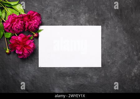 Biglietto d'auguri con fiori di peonie rosse e carta bianca bianca su sfondo scuro, piatto, vista dall'alto. Concetto di vacanza con spazio di copia per il testo per il vostro Foto Stock