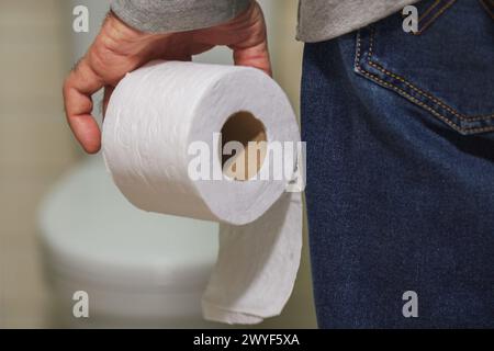 La mano di un uomo anziano che tiene un rotolo di carta igienica davanti al bagno. Foto Stock