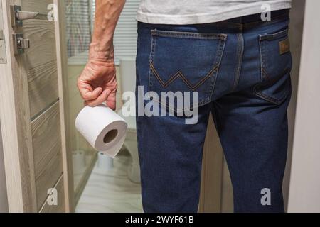 La mano di un uomo anziano che tiene un rotolo di carta igienica davanti al bagno. Foto Stock