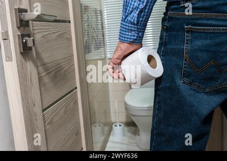 La mano di un uomo anziano che tiene un rotolo di carta igienica davanti al bagno. Foto Stock