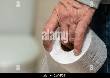 La mano di un uomo anziano che tiene un rotolo di carta igienica davanti al bagno. Foto Stock