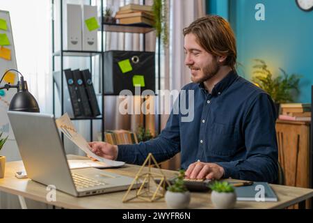 Giovane lavoratore d'ufficio che lavora con programmi e grafica finanziaria utilizzando calcolatrice e laptop seduti alla scrivania dell'ufficio domestico. Buon uomo d'affari libero professionista confronta e analizza grafici grafici grafici grafici grafici grafici e grafici. Foto Stock