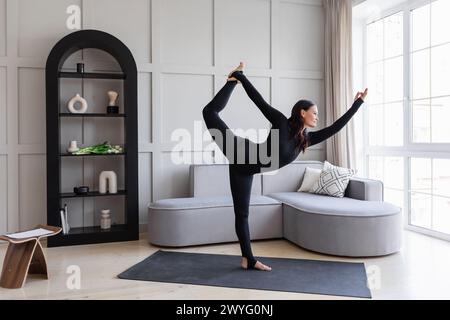 Attraente donna bruna che esegue l'esercizio Natarajasana a casa, danza in posa, pratica yoga in tute sportive nere in piedi su un tappetino, pieno Foto Stock