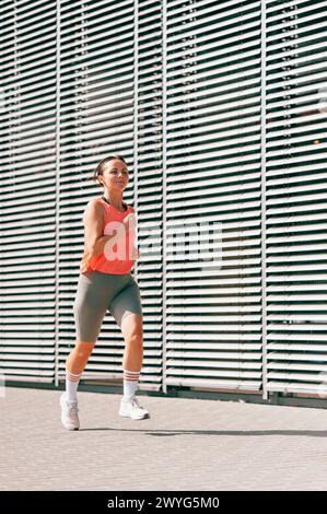 La foto integrale di una donna di mezza età in forma corre all'aperto in una giornata di sole. Parete metallica come sfondo. Foto Stock