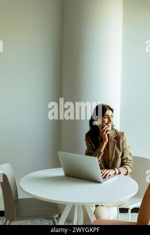 Una donna siede a un tavolo bianco in un ufficio illuminato dal sole, perso nel pensiero, con un portatile e una pianta verde accanto a lei. Foto Stock