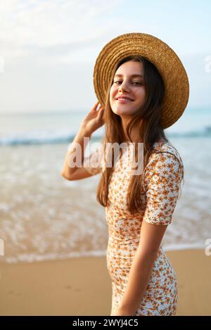 Una giovane donna sorridente con un abito floreale e un cappello di paglia si erge sulla spiaggia sabbiosa. La donna alla moda ama l'estate al tramonto sul mare. Atmosfera vintage, viaggi Foto Stock