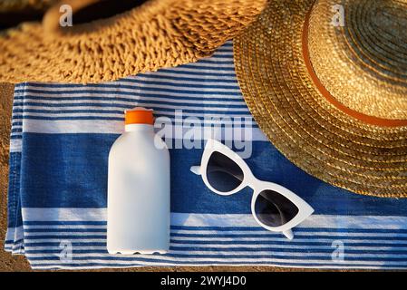 Foto in alto degli indispensabili estivi sulla spiaggia sabbiosa, bottiglia di crema solare bianca accanto a eleganti occhiali da sole, oltre a un asciugamano a righe sotto il cappello in tessuto per un look elegante Foto Stock