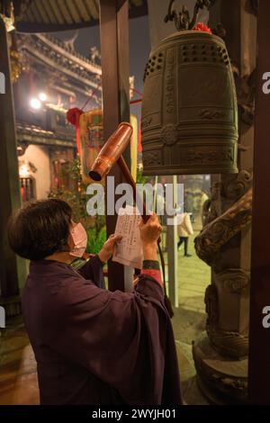 Donna abbigliamento tradizionale che suona una grande campana in una cerimonia del tempio Baoan, durante il capodanno cinese Foto Stock