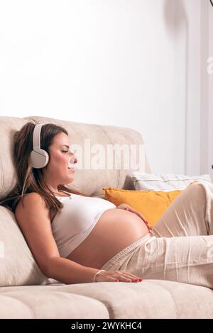 Giovane donna incinta che ascolta la musica con le cuffie mentre si rilassa sul divano di casa. Foto di alta qualità Foto Stock