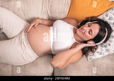 Giovane donna incinta che ascolta la musica con le cuffie mentre si riposa sul divano di casa. Foto Stock