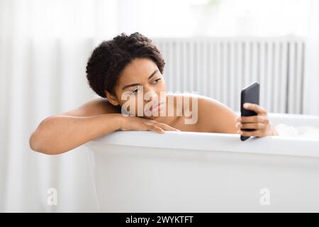 Pensiva donna afroamericana nella vasca da bagno con telefono Foto Stock