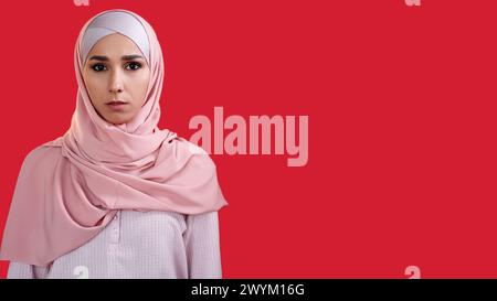 Ritratto femminile. Volto curioso. Bella donna sicura di sé con foulard hijab di colore rosa isolato su sfondo rosso vuoto. Foto Stock
