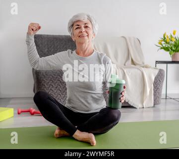 Una donna anziana allegra guarda nella fotocamera e mostra il suo Bicep, incarnando uno stile di vita attivo e gli allenamenti domestici Foto Stock